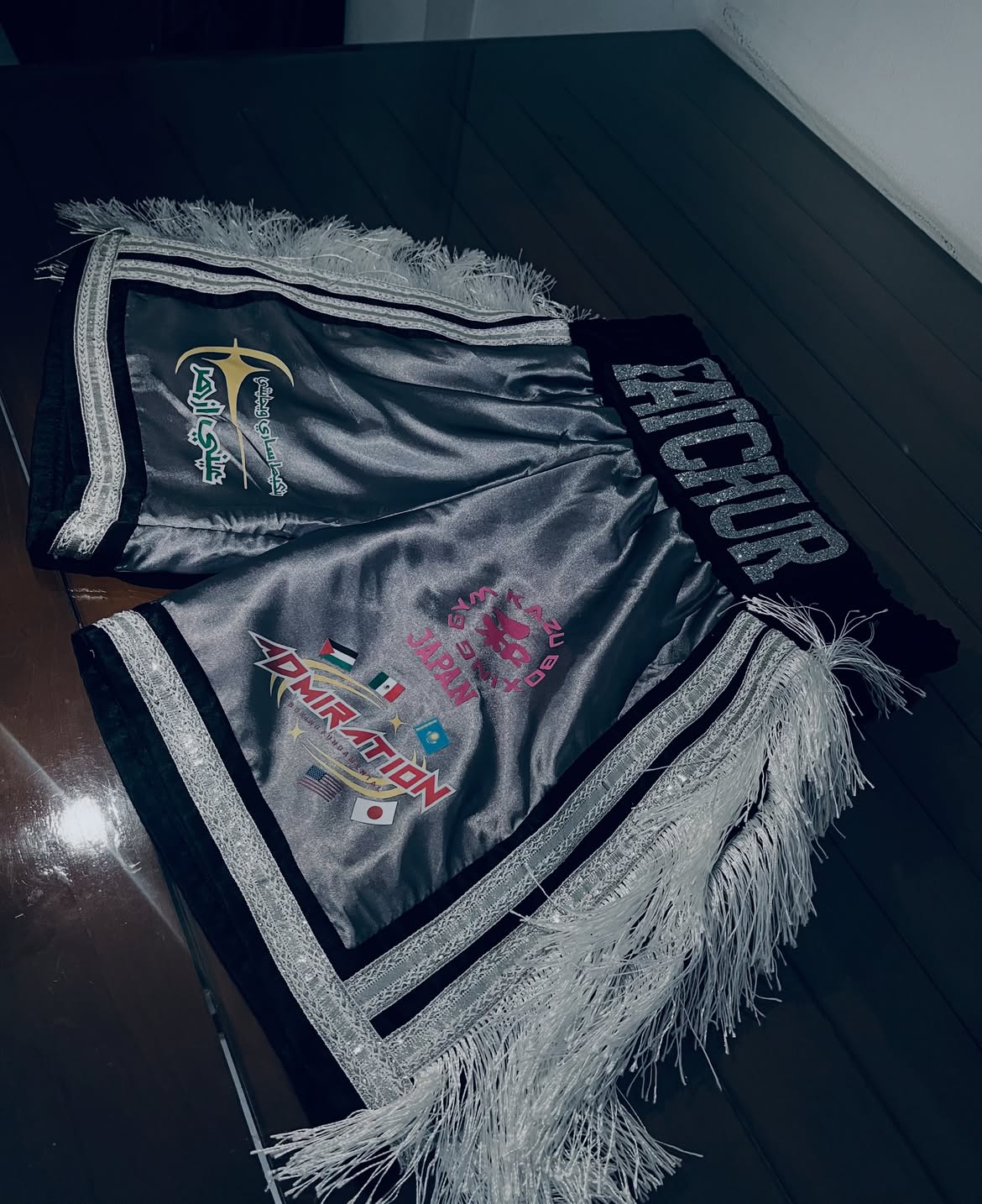Silver Khodir boxing trunks with a black waistband, white fringe and custom patches.