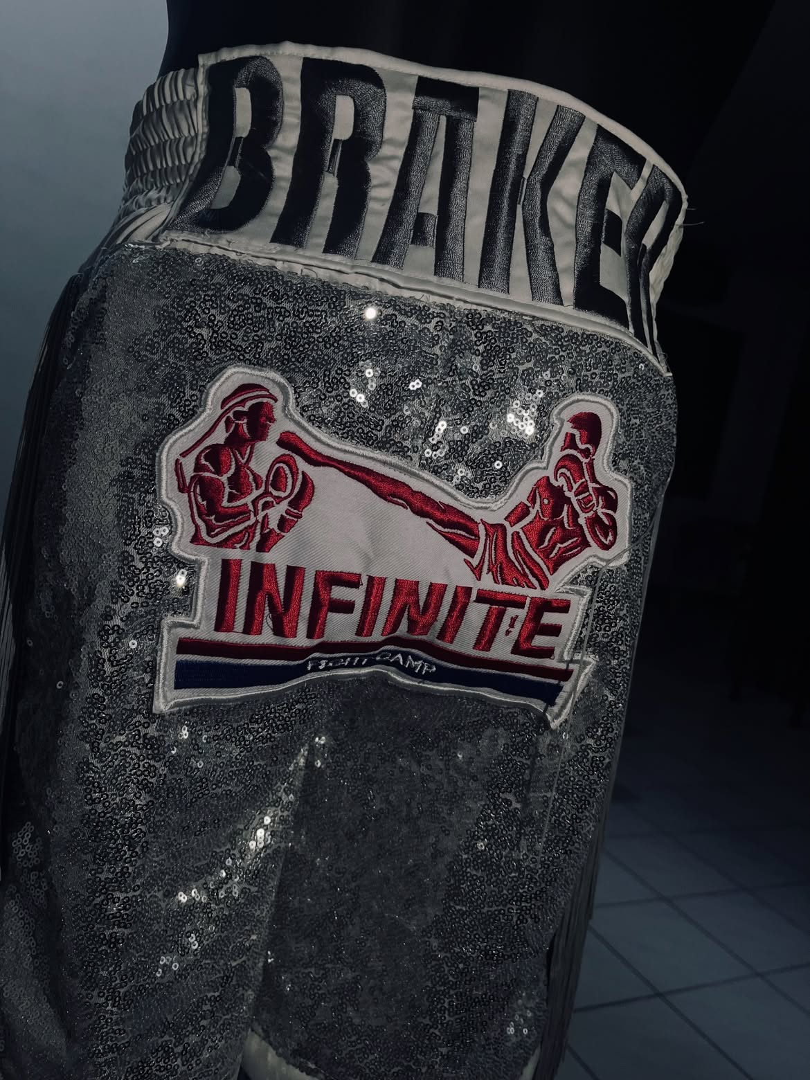 Close-up of the Silver Infinite boxing trunks showing the sequin body, embroidered kickboxing artwork and waistband lettering.