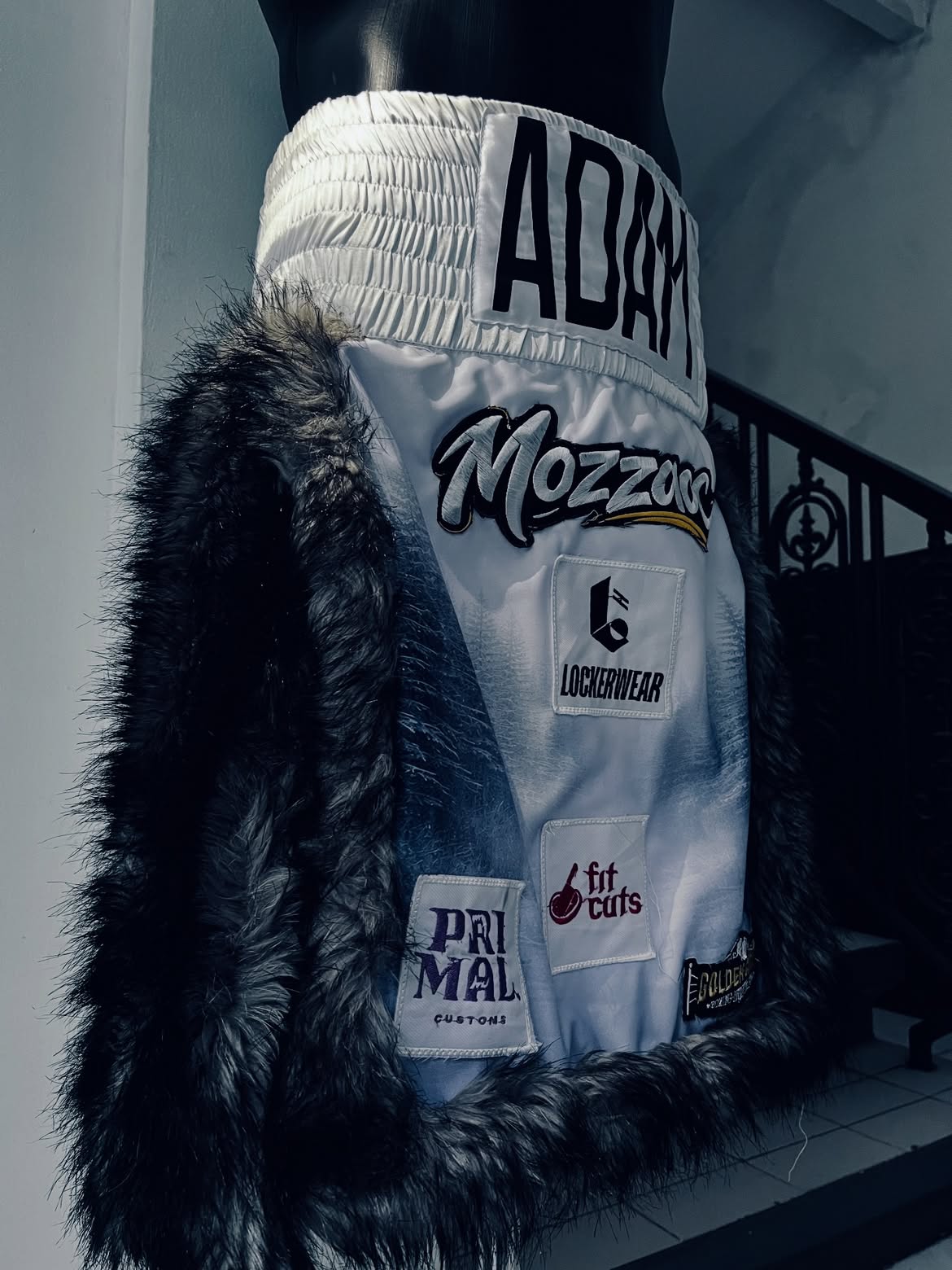 White Adam Maulana boxing trunks with black faux-fur trim and custom patches.